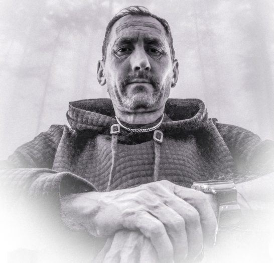 A man in a hoodie sits with hands clasped, surrounded by a misty, forested background.