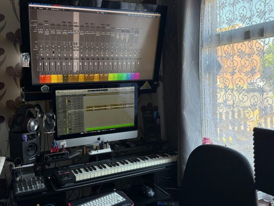 A music production workspace with two monitors, a keyboard, and natural light from a window.